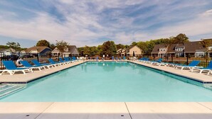 Uma piscina retangular com cadeiras de descanso azuis de cada lado, cercada por uma grade, e casas ao fundo.