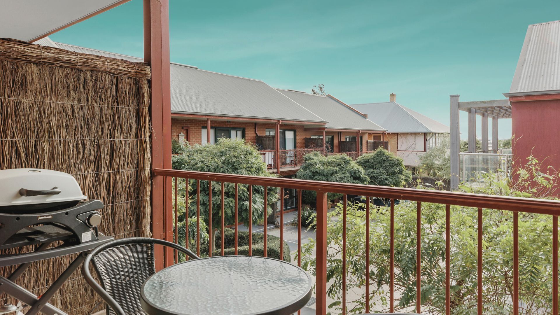 Ein Balkon mit Grill, Korbsessel und Tisch, mit Blick auf eine Wohngegend mit Häusern und Grünflächen.