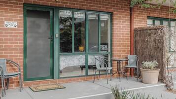 A brick building with a green-framed door, two chairs, a small table, and a potted plant.