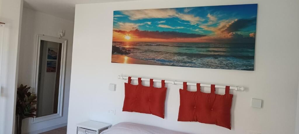 Un dormitorio con una cama, una cortina roja, un cuadro de un atardecer y una ventana.