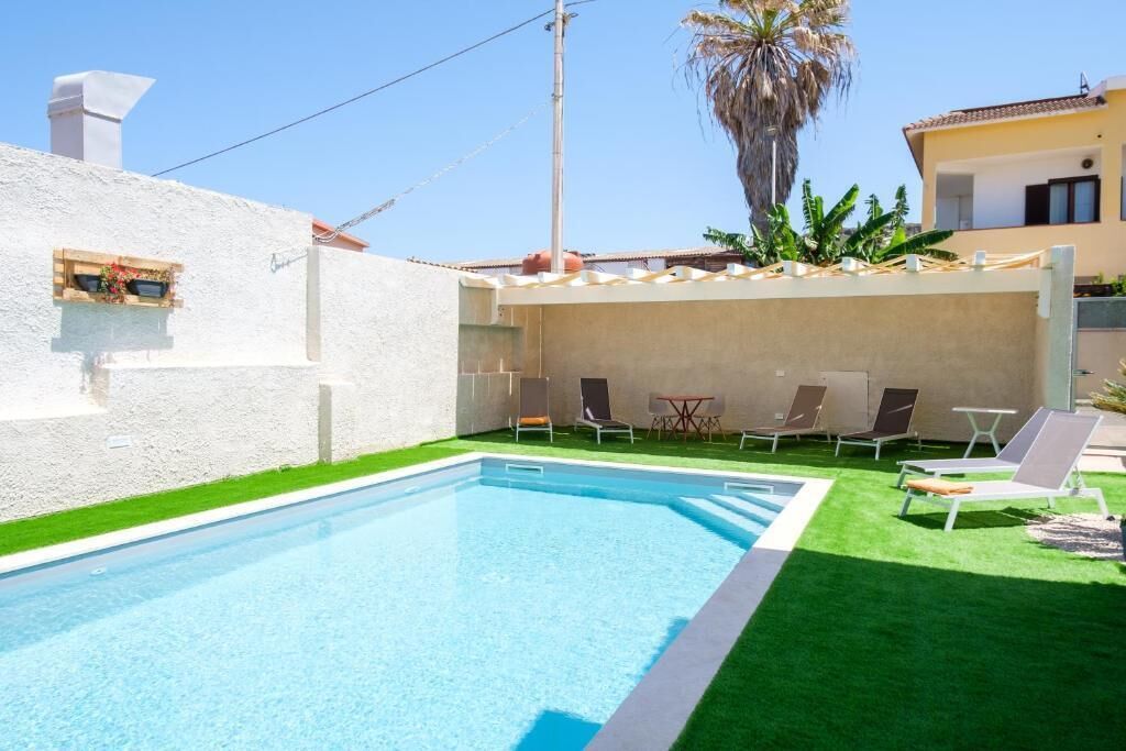 A swimming pool with lounge chairs and a small table, surrounded by a white wall and green grass.