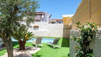 A pool area with two lounge chairs, a palm tree, and a white wall.