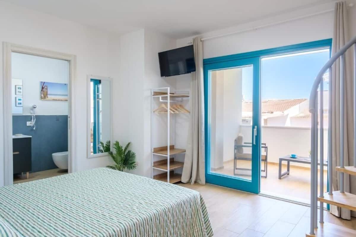 Habitación de hotel con cama, baño, televisión, balcón con mesa y vista a la ciudad.