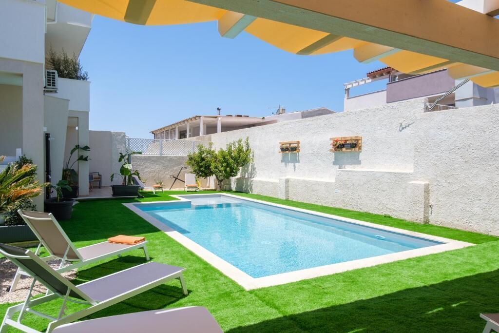 A swimming pool with a white pool edge, surrounded by artificial grass, and a white wall with windows.