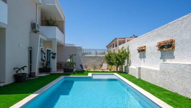 Piscina com água azul cristalina, rodeada por um edifício branco com varandas e um gramado verde.