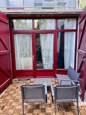 Eine Terrasse mit zwei Stühlen und einem Tisch, eine rote Tür mit Fensterfeldern und ein weißer Vorhang.