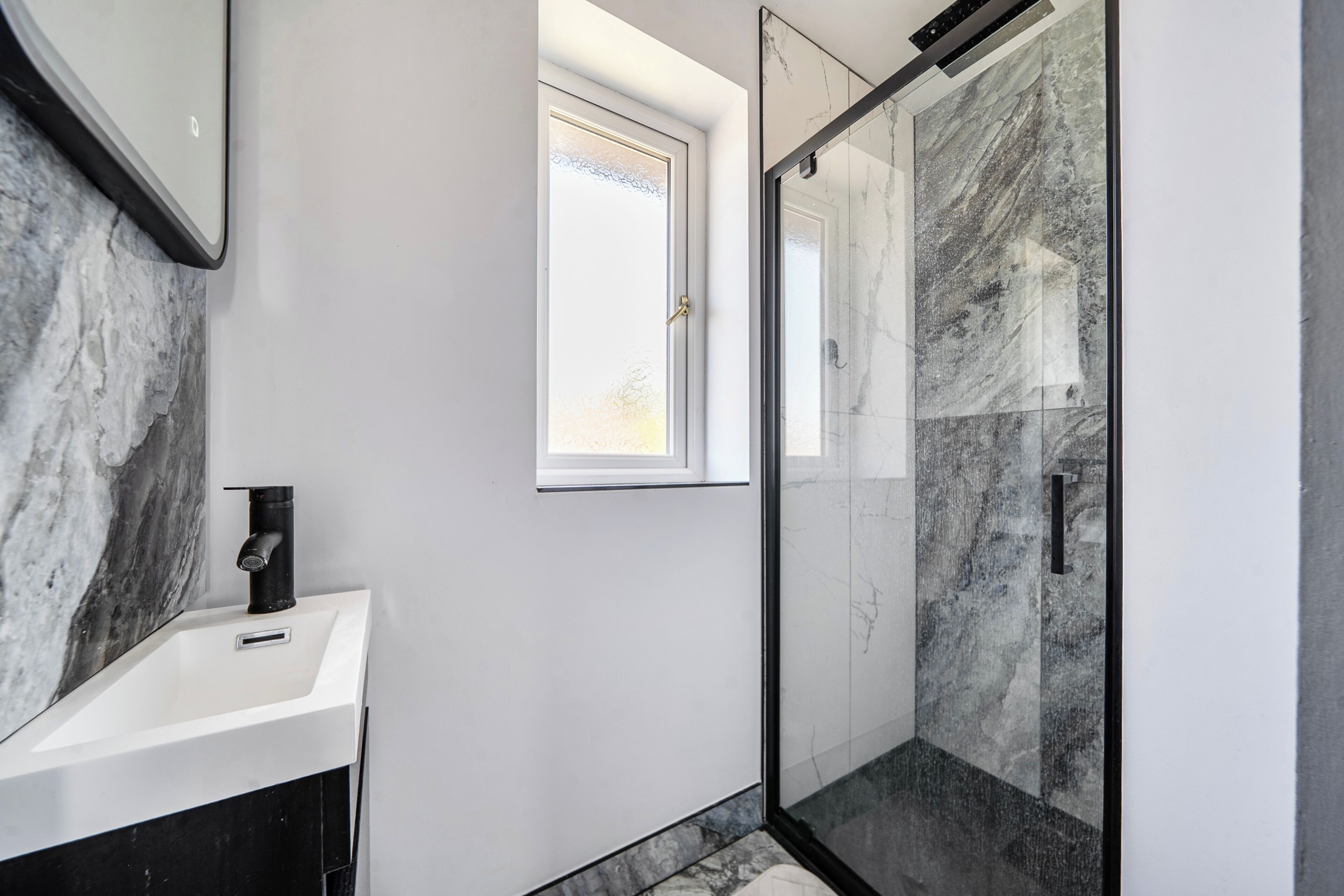 A bathroom with a white sink, a black faucet, a glass shower enclosure, and a window.