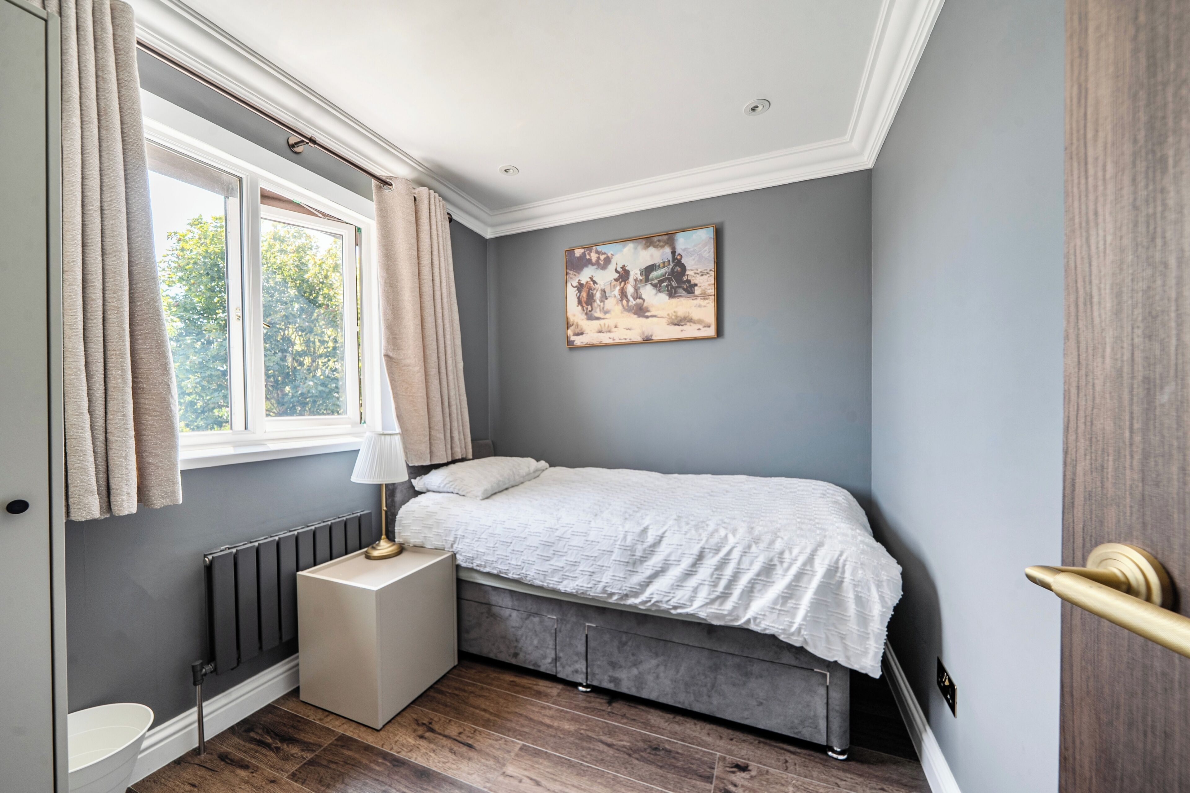 A bedroom with a bed, a radiator, a window with curtains, a bedside table with a lamp, and a painting on the wall.
