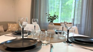 Ein Esstisch ist gedeckt mit Tellern, Gläsern und Besteck, im Hintergrund ist ein Fenster mit Blick auf Grünpflanzen zu sehen.