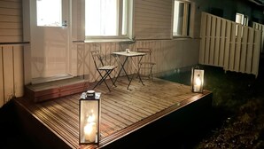 Eine Holzdielen-Terrasse mit Tisch, Stühlen, zwei Fenstern und einer Laterne.