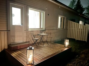 Eine Holzdielen-Terrasse mit Tisch, Stühlen, zwei Fenstern und einer Laterne.