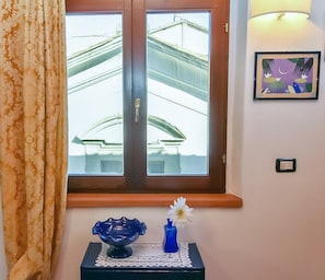 Quarto com janela, luminária fixada na parede, quadro emoldurado e pequena mesa com um vaso e uma tigela decorativa.