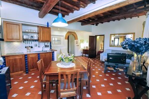 Uma área de refeições com mesa e cadeiras de madeira, uma luminária pendente azul e uma cozinha com armários de madeira e painel de azulejos.