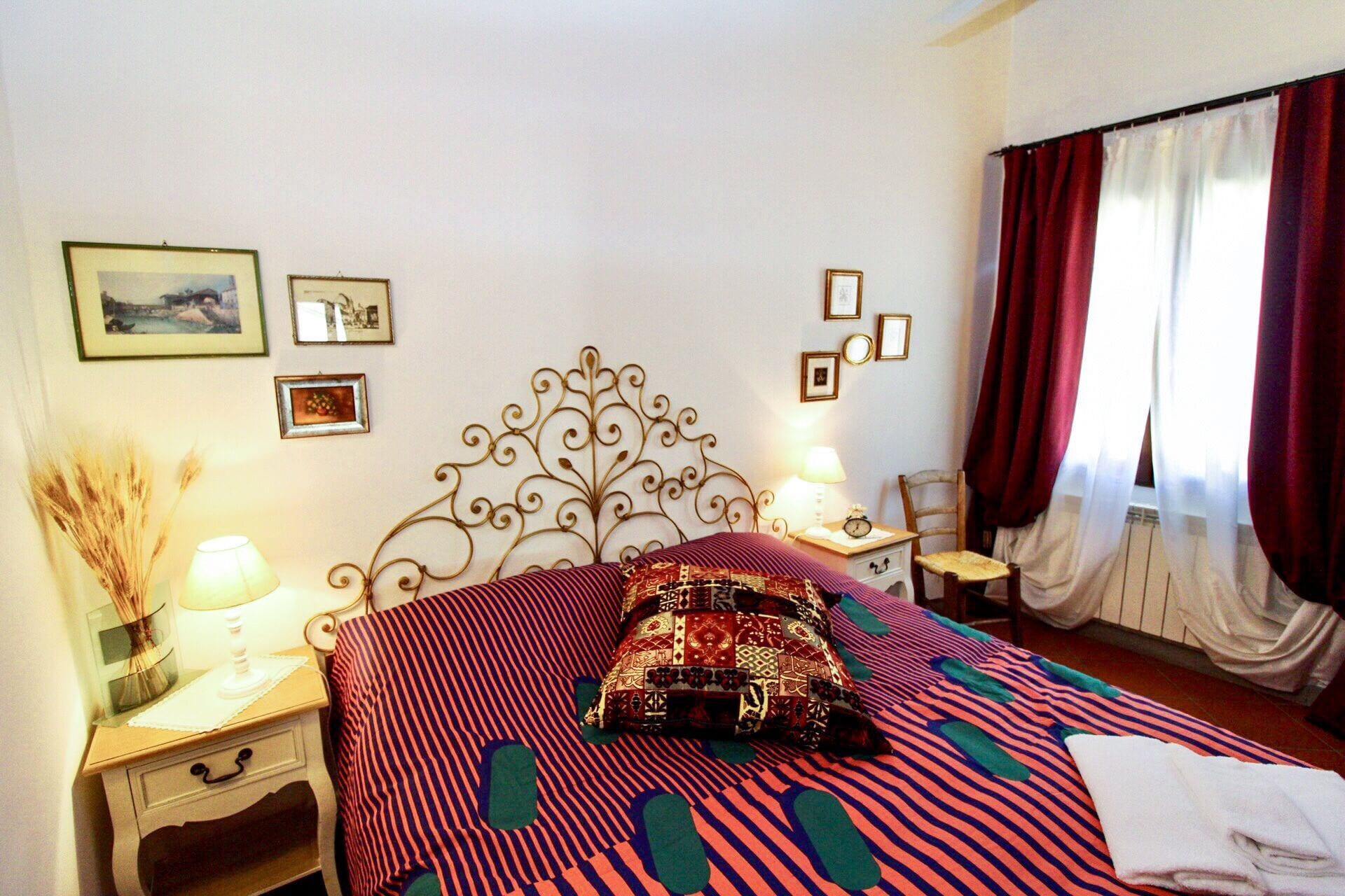 Una habitación de hotel con una cama, mesitas de noche, una silla, un ventanal con cortinas y adornos en la pared.