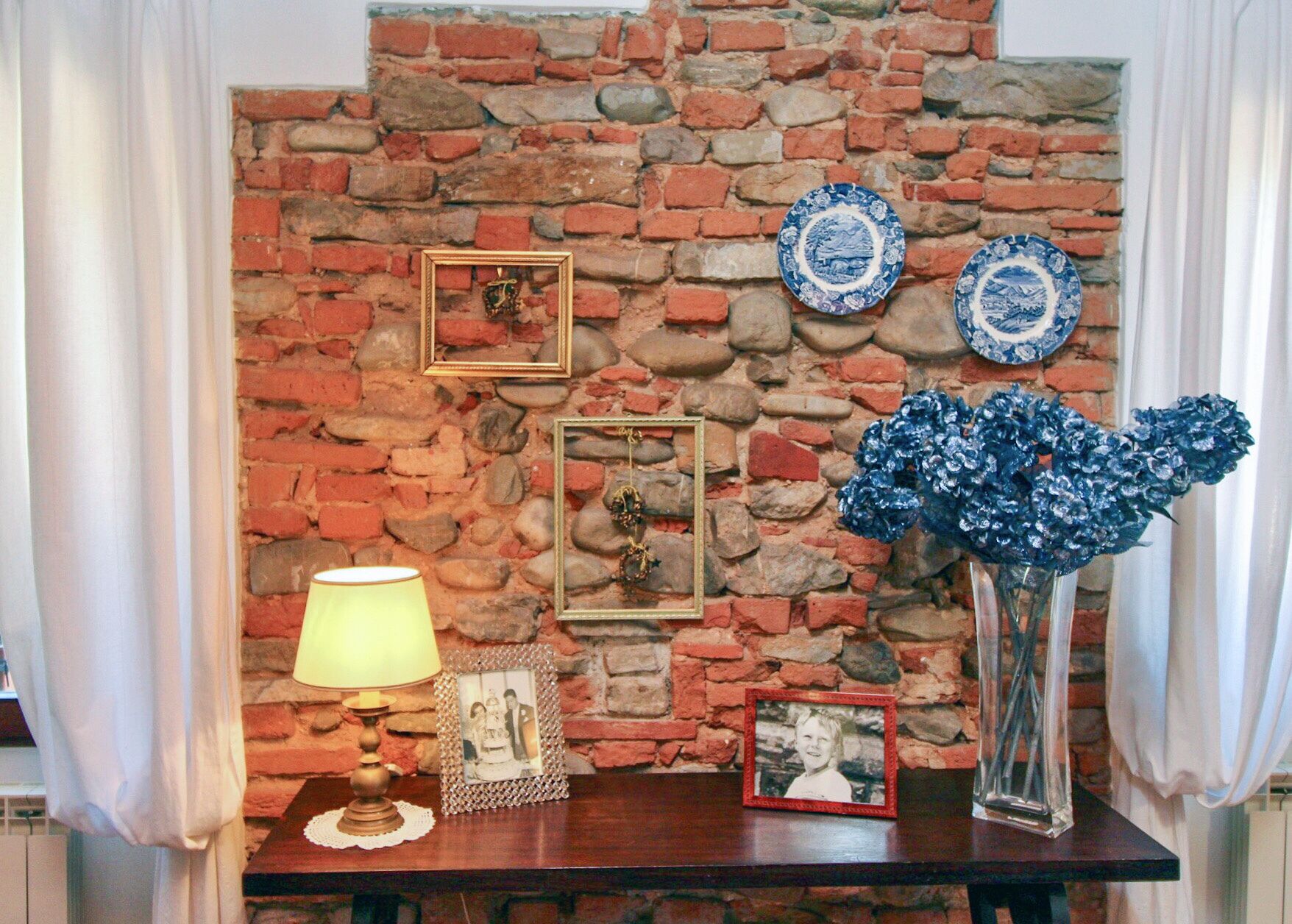 Une pièce avec un mur en briques, une table en bois, une lampe, des photos encadrées et des assiettes décoratives.