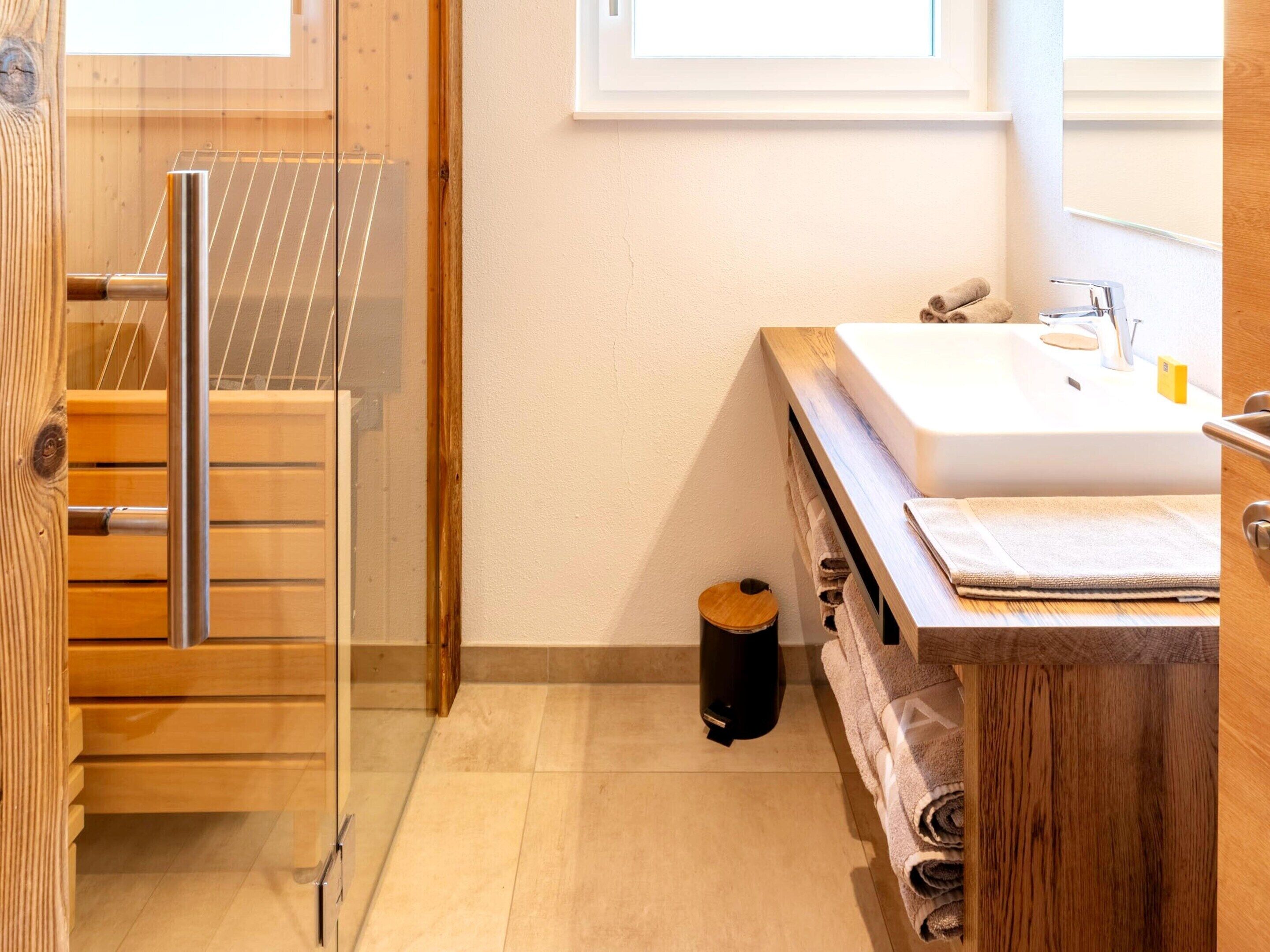 Ein Badezimmer mit Holztür, Duschkabine aus Glas, Waschbecken mit Handtuch und einem Mülleimer.