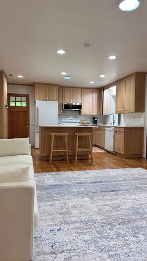 A modern kitchen with wooden cabinets, a white refrigerator, a stove, and a microwave. There are two wooden stools at the kitchen island. - Tranquil Nature Retreat: Enjoy this Beautiful Home surrounded by Trees & Flowers (Winston-Salem)