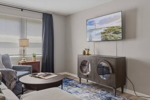 A living room with a sofa, a round coffee table, a cabinet with circular cutouts, a TV mounted on the wall, and a window with blinds. - Modern Corning Unit | Pet OK, Walk to CMOG/Gaffer (Corning)