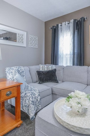 A modern living room with a grey sectional sofa, a wooden side table, and a round coffee table with a white floral centerpiece. - Sun-Filled Upper Gem | Near LECOM & Elmira College (Elmira)