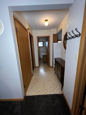 A hallway with a carpeted floor, a wooden door, a wall-mounted light fixture, and a coat rack.