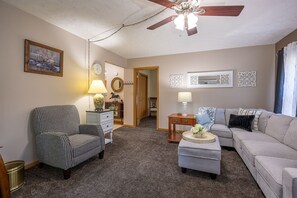 Un salon comprenant un fauteuil à motif pied-de-poule, un canapé gris, une table basse avec un vase, une table d’appoint en bois, une lampe, une horloge et un ventilateur de plafond.