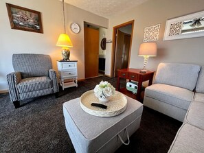 Un salon avec un fauteuil à motif pied-de-poule, un canapé gris, une table basse ornée d’un vase de fleurs, une lampe et une horloge au mur.