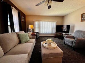 Un salon comprenant un canapé, un pouf et un fauteuil à motifs, une télévision et un ventilateur de plafond.