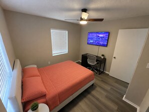 A bedroom with a bed, a desk, a chair, a television, and a ceiling fan.