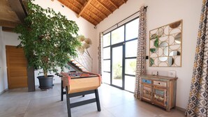 Una habitación amplia con una mesa de fútbol, una planta grande en maceta, un aparador de madera y un ventanal con cortinas.