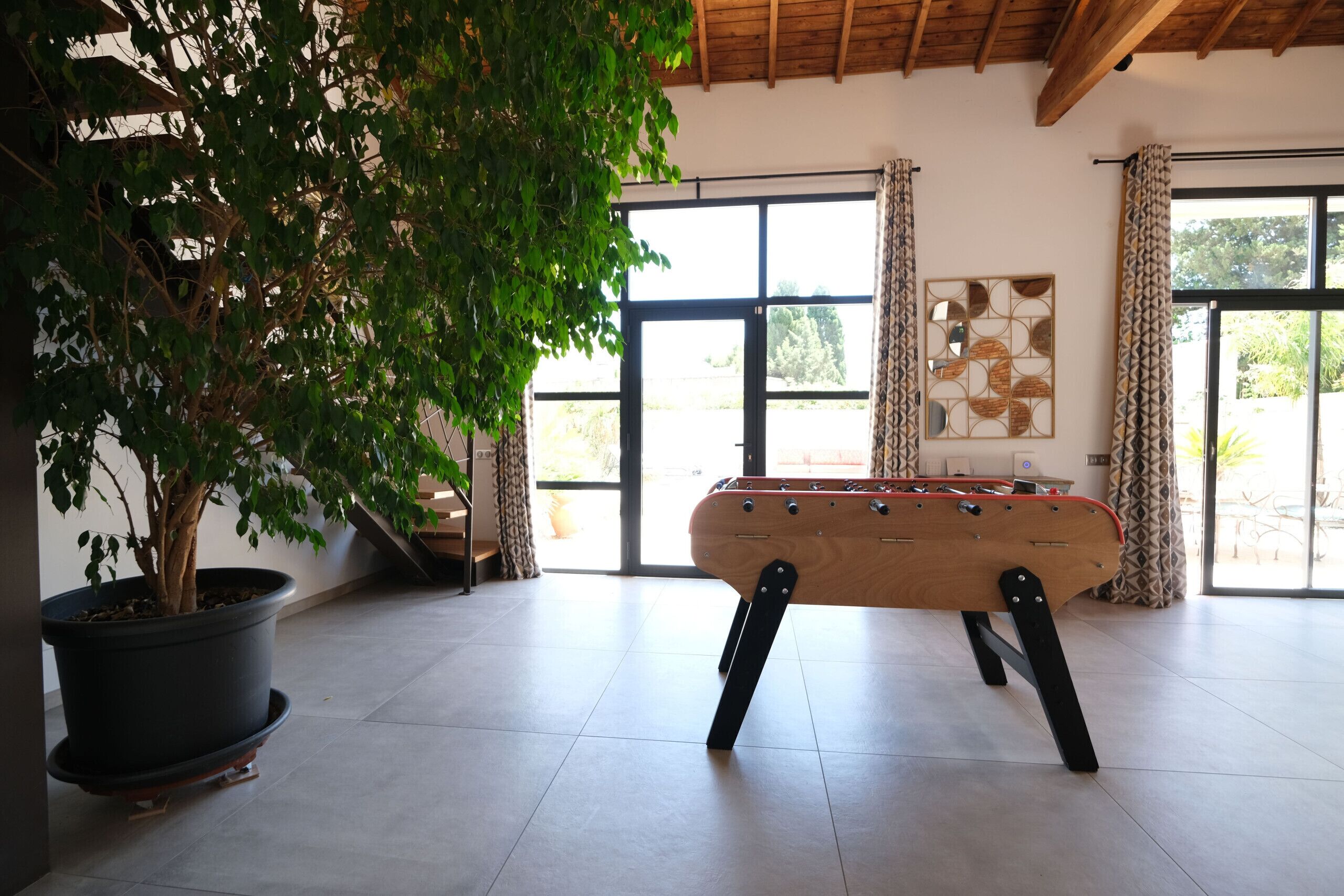 A room with a foosball table, a potted plant, and a large window with curtains.
