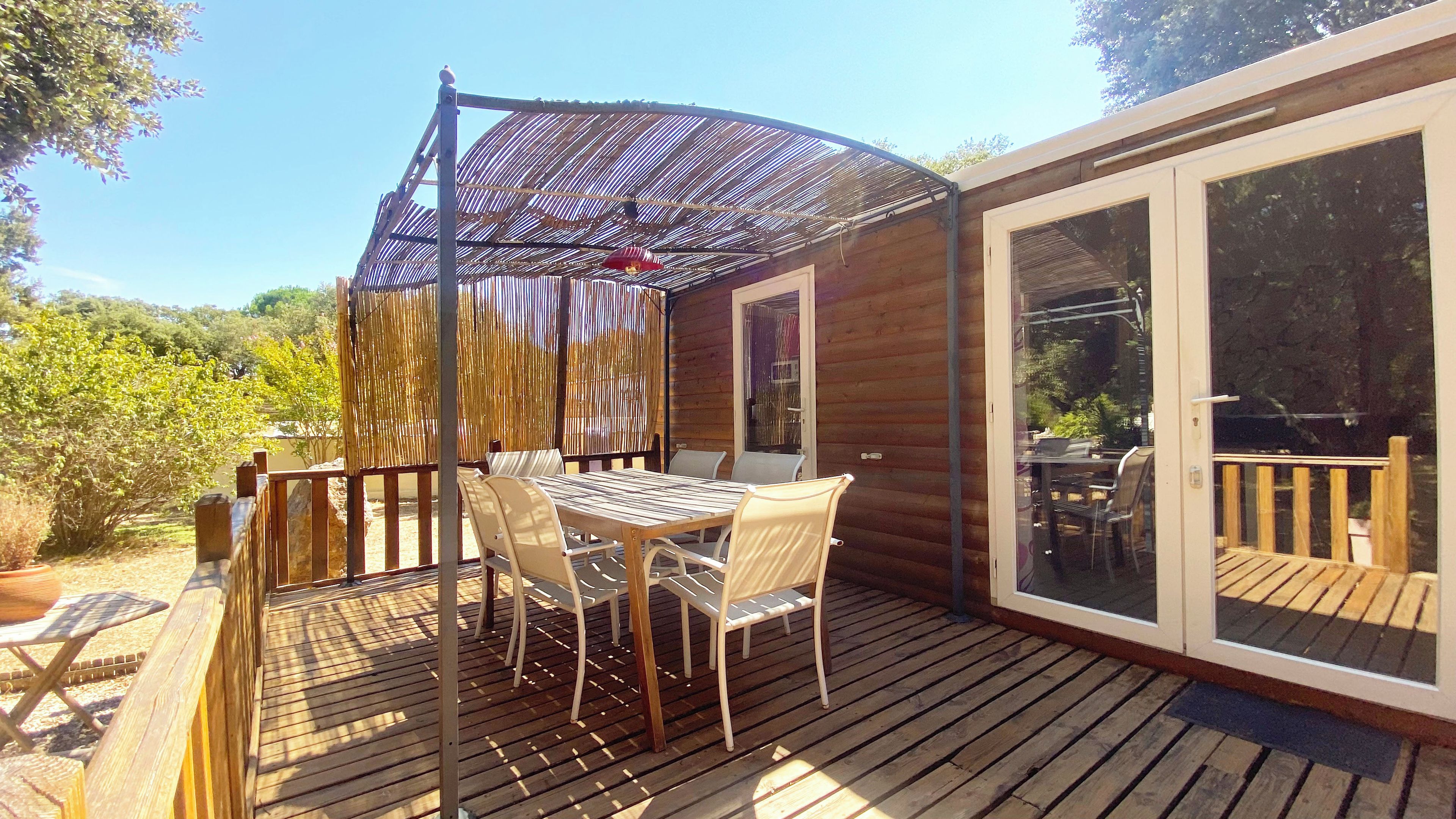 Eine Holzterrasse mit Tisch und Stühlen, einem überdachten Sitzbereich und einer Glastür zum Außenbereich.