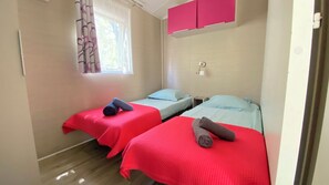 A room with two beds, each with a red bedspread and a pillow. There is a window with curtains, a pink cabinet above the beds, and a door on the right.