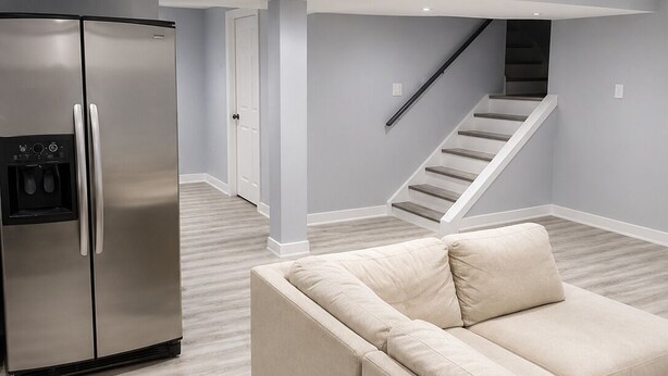 A modern living room with a sectional sofa, a stainless steel refrigerator, and a staircase leading to an upper level.