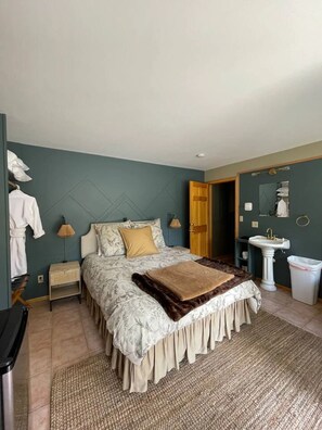 Un dormitorio con cama, mesitas de noche, lámpara, espejo, lavamanos y un cubo de basura.