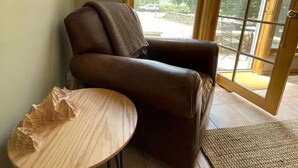 Ein Zimmer mit braunem Ledersessel, einem kleinen runden Tisch, einem an der Wand montierten Fernseher und einem großen Fenster mit Blick ins Freie.