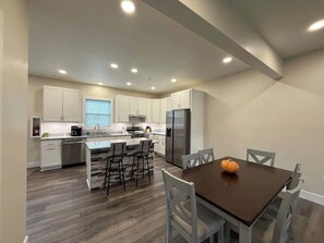 A modern kitchen with white cabinets, a central island with bar stools, a dining table with chairs, and a refrigerator. - Willow Creek Townhouse (Aliquippa)
