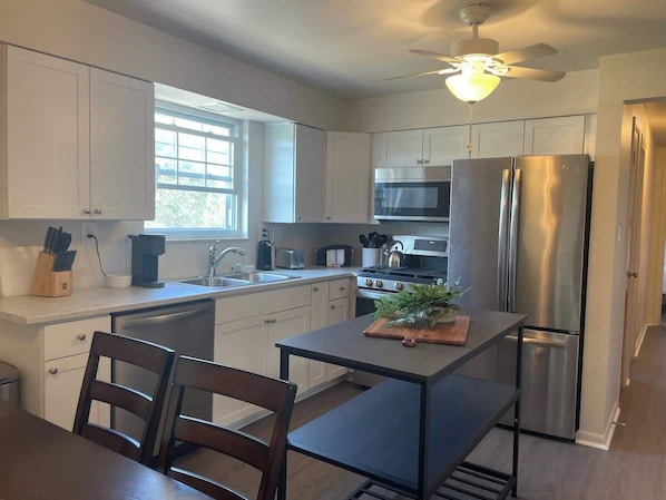 A modern kitchen with white cabinets, stainless steel appliances, a dining table with chairs, and a ceiling fan.