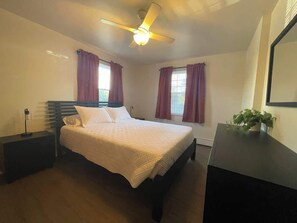 A bedroom with a bed, a ceiling fan, a nightstand, a dresser, and a window with curtains.