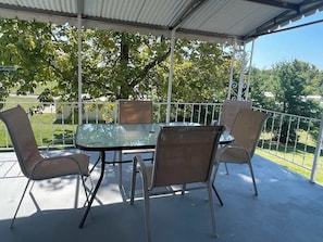 A covered patio with a glass table and four chairs, surrounded by a white fence and trees.