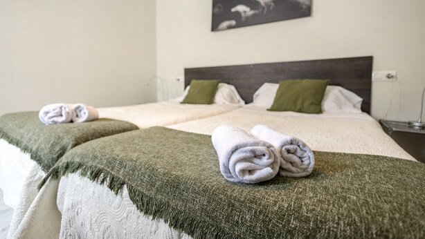 A hotel room with two beds, each with a green throw blanket and a rolled towel. There is a painting on the wall above the beds. - GuestReady - Atico Luna Puerto (El Puerto de Santa María)