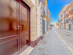 A street view with a closed brown door, a tiled sidewalk, and buildings with balconies and windows. - GuestReady - Atico Luna Puerto (El Puerto de Santa María)