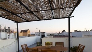 A rooftop patio with a wooden table and chairs, a potted plant, and a view of buildings in the background. - GuestReady - Atico Luna Puerto (El Puerto de Santa María)