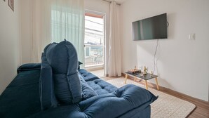 Uma sala de estar com sofá azul, televisão fixada na parede, mesinha com objetos decorativos e vista pela janela com cortinas.