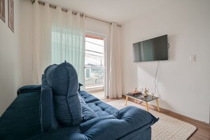 Uma sala de estar com sofá azul, televisão fixada na parede, mesinha com objetos decorativos e vista pela janela com cortinas.