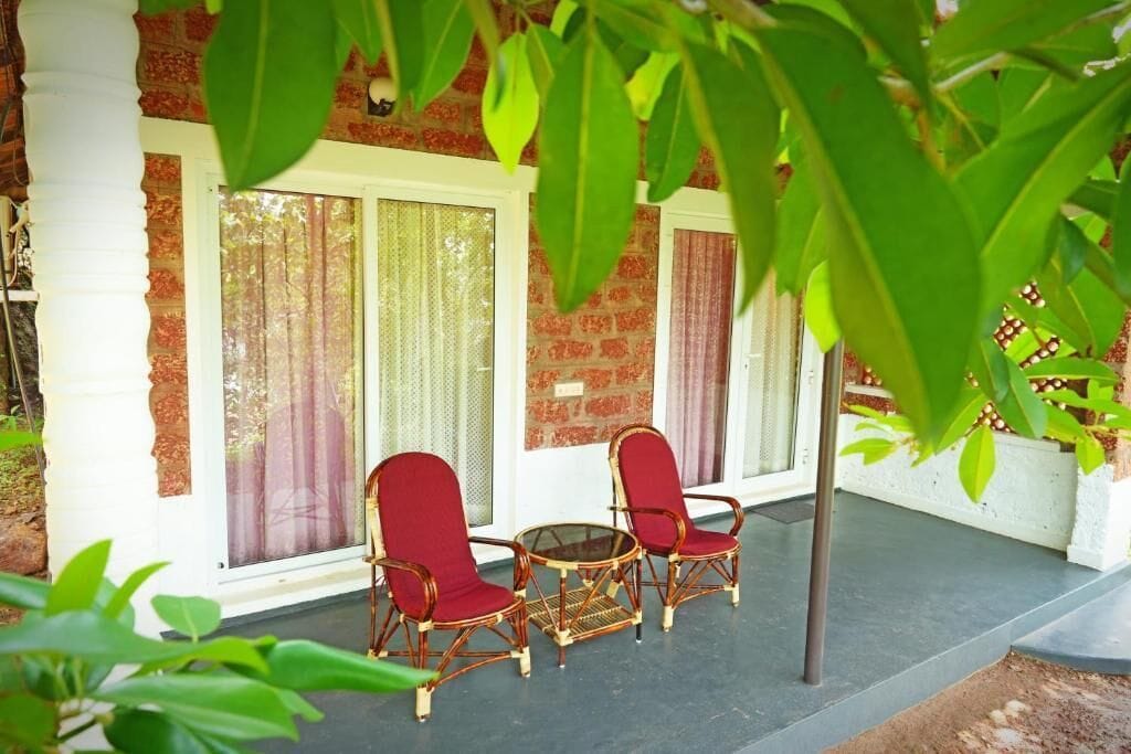Un espacio al aire libre techado con sillas de mimbre y una mesa de vidrio, junto a un edificio con ventanas amplias y una pared de ladrillo.