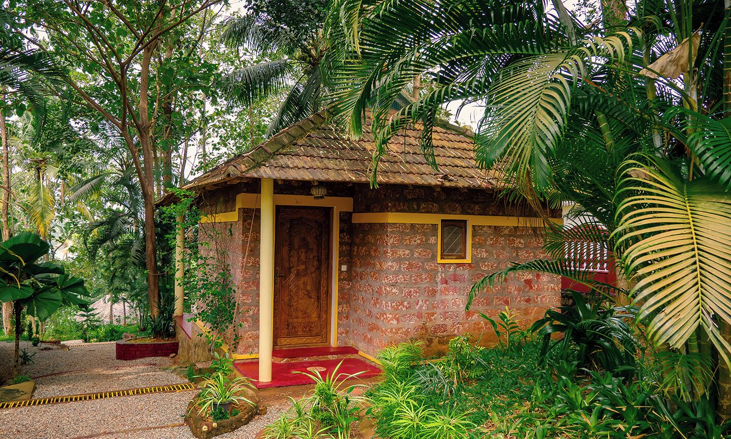 A small, rustic hut with a thatched roof, surrounded by lush greenery and a gravel path.