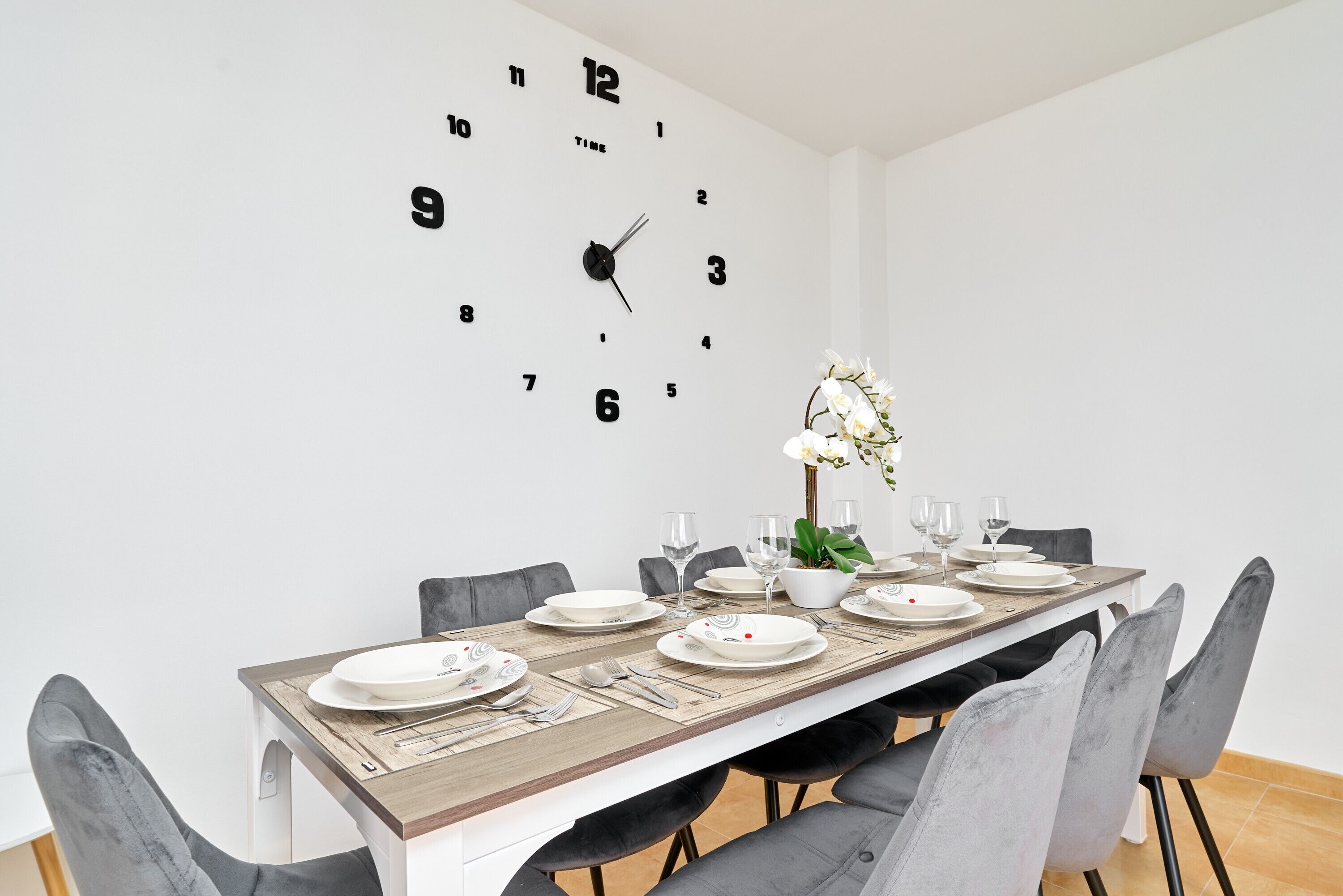 Une salle à manger avec une longue table, des chaises grises, une horloge au mur et une plante en pot.