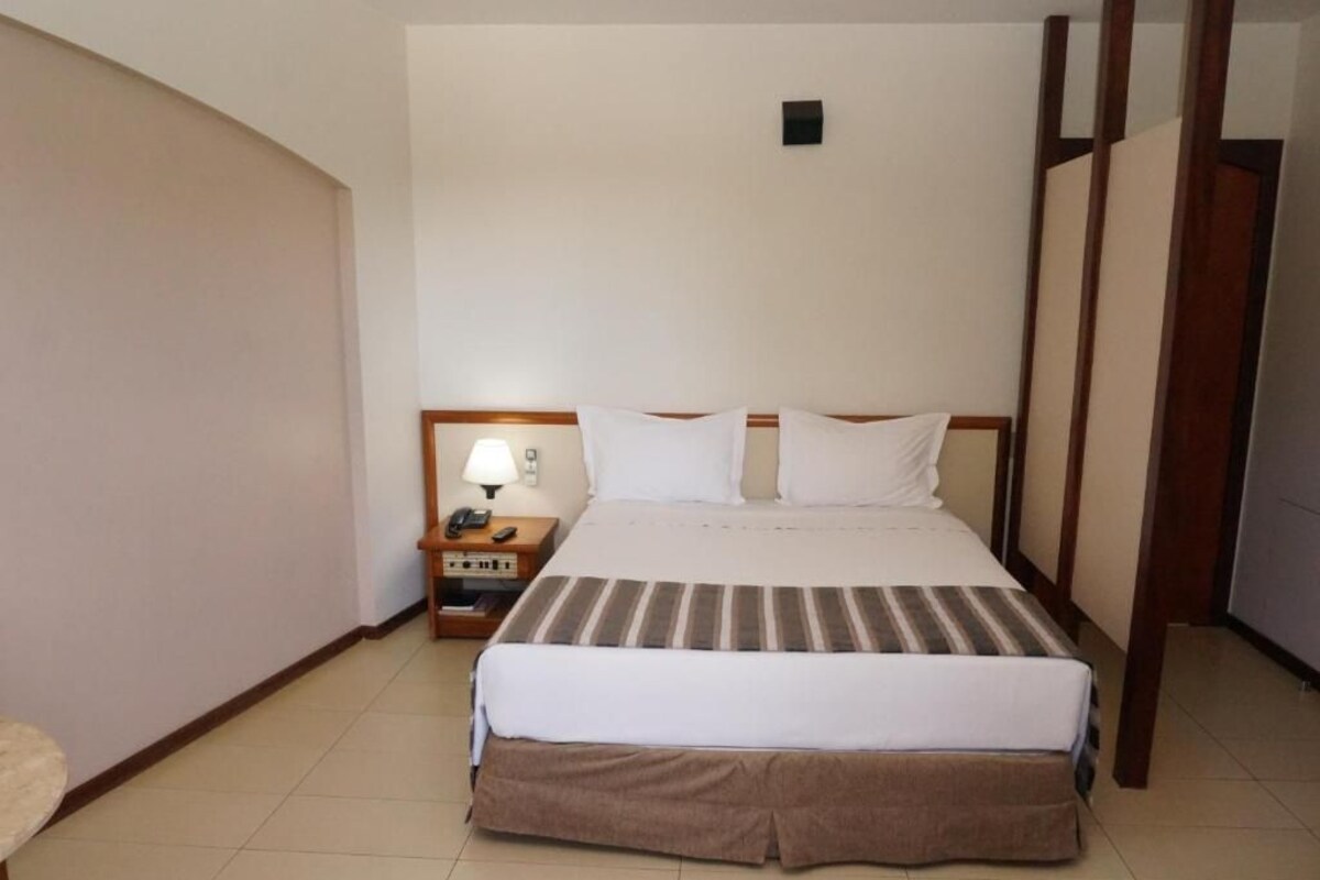 A hotel room with a large bed, a bedside table with a lamp, and a small cabinet.