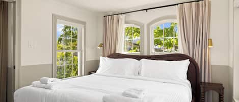 Una cama bien tendida con sábanas y toallas blancas, cabecera de madera y dos ventanas con cortinas.