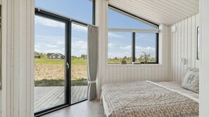 Un dormitorio con un amplio ventanal corredizo, una cama con colcha estampada y una ventana con vistas a una casa y un campo.